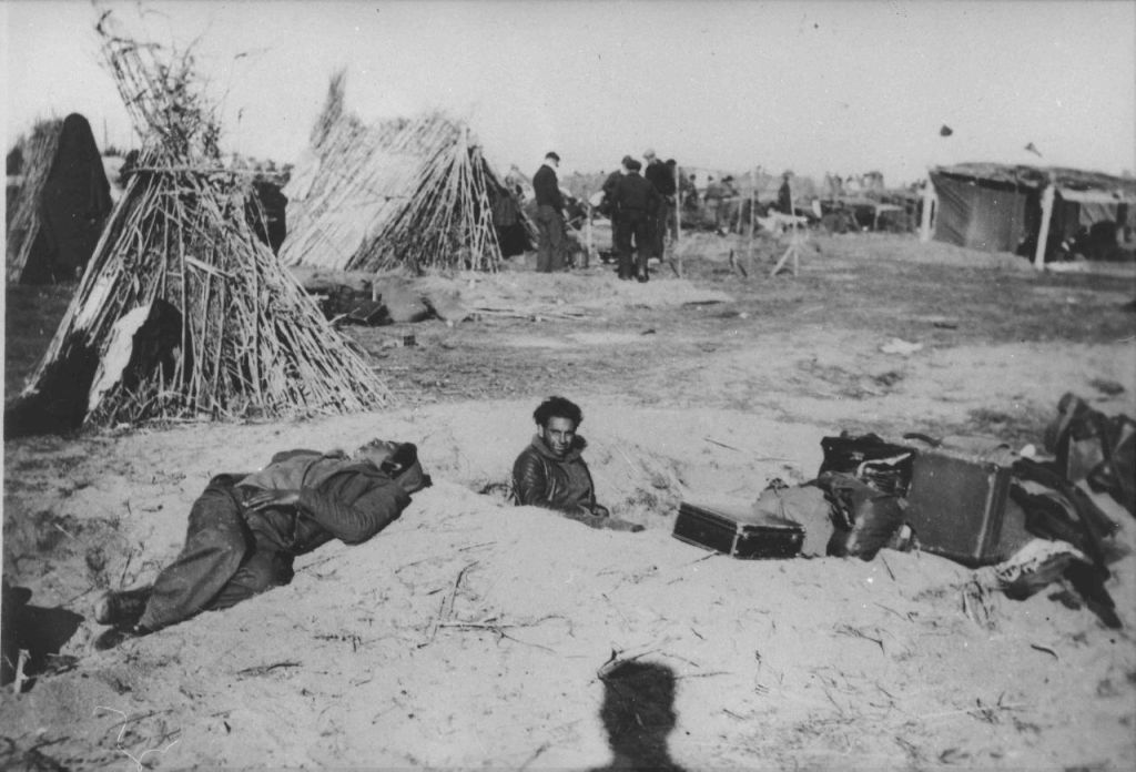 Black and white photo taken from inside a refugee camp. In the background are a few very rough reed shelters, some fencing, and a few men. In the foreground, two men lie or sit in shallow holes dug into the sand. To the right there is a pile of suitcases and other luggage.